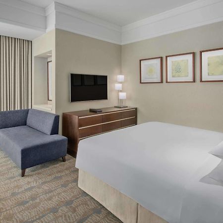 Jabal Omar Hyatt Regency Makkah Hotel Mekka Eksteriør billede The photo shows a modern hotel room featuring a neatly made bed with white linens, a small blue couch to the side, and a wooden dresser with a television mounted on the wall above it. There are two framed pieces of art on the wall near the bed, and t