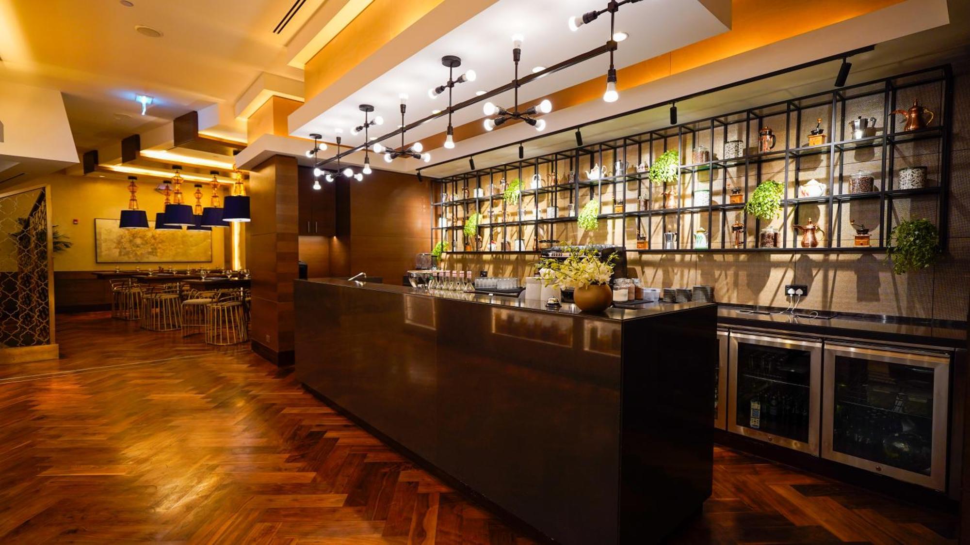 Jabal Omar Hyatt Regency Makkah Hotel Mekka Eksteriør billede The photo shows a modern bar area in a restaurant. The bar features a sleek, dark countertop and is backed by a decorative arrangement of shelves that holds various bottles and plants. The lighting is warm and stylish, with multiple hanging pendant l
