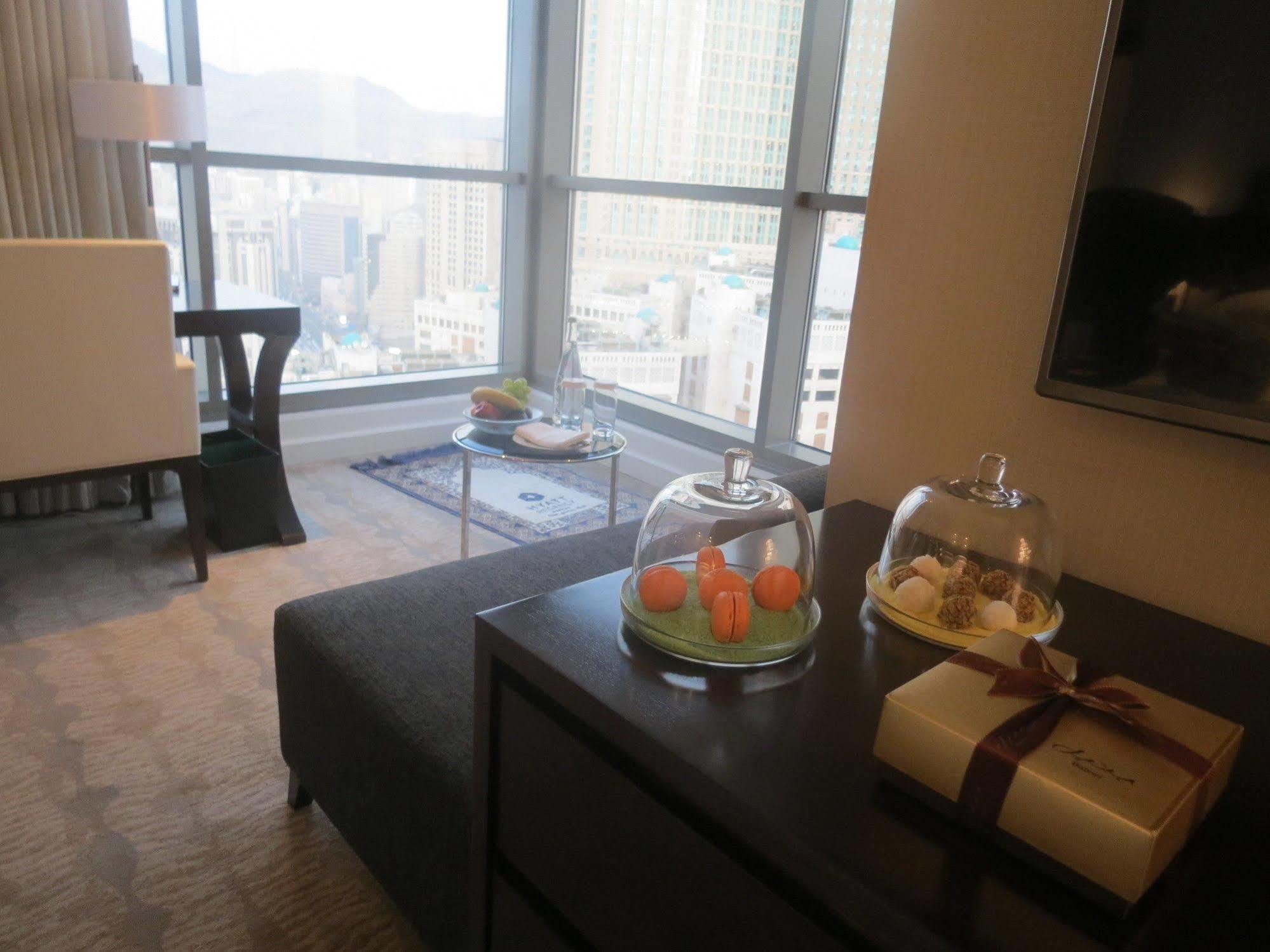 Jabal Omar Hyatt Regency Makkah Hotel Mekka Eksteriør billede The photo shows a modern hotel room or a stylish living space with large windows offering a view of the city. In the foreground, there is a dark sofa and a low table with covered trays displaying what appears to be small desserts or delicacies. To th