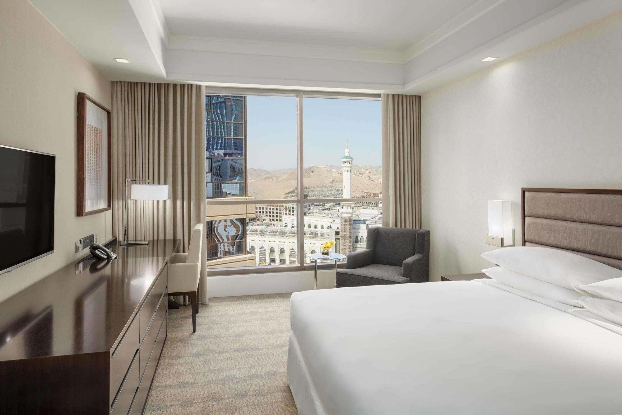 Jabal Omar Hyatt Regency Makkah Hotel Mekka Eksteriør billede The photo shows a modern hotel room featuring a large window that provides a view of an outdoor landscape, likely part of a city or landmark. The room includes a bed with white linens, a side table with a lamp, and a small seating area with a chair a