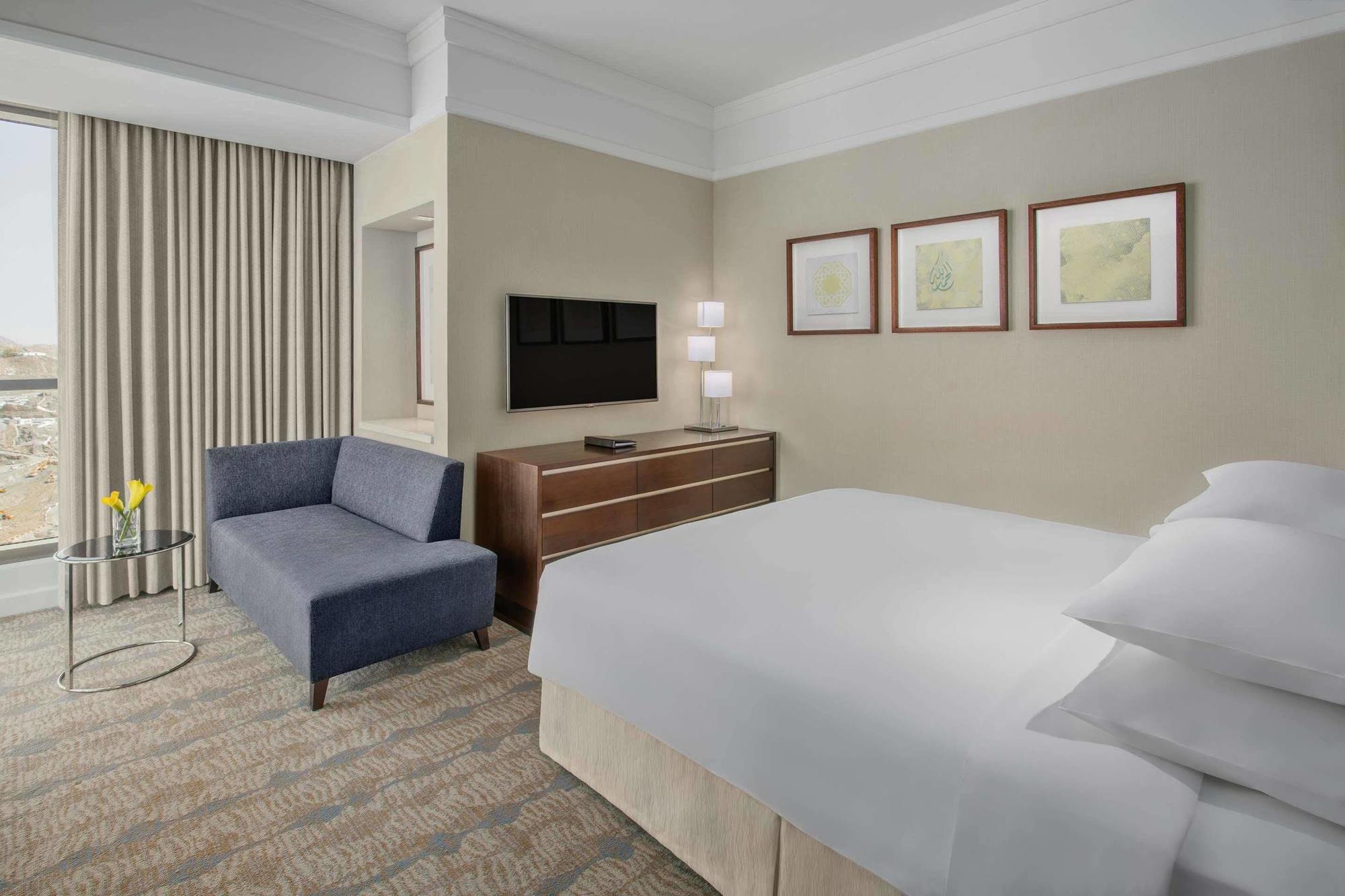 Jabal Omar Hyatt Regency Makkah Hotel Mekka Eksteriør billede The photo shows a modern hotel room featuring a neatly made bed with white linens, a small blue couch to the side, and a wooden dresser with a television mounted on the wall above it. There are two framed pieces of art on the wall near the bed, and t