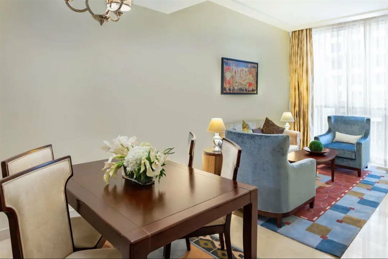 Jabal Omar Hyatt Regency Makkah Hotel Mekka Eksteriør billede The photo shows an elegantly furnished living space. On the left, there's a wooden dining table with four chairs, decorated with a vase of white flowers. To the right, there are two comfortable blue armchairs and a small side table, each adorned with
